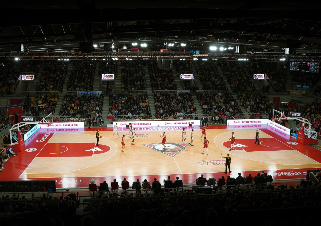 A basketball arena mid-game, LED perimeter boards running sponsor banners around the court